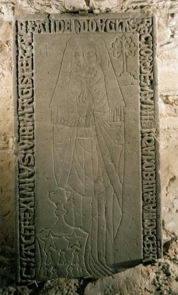 Colour photo of a slab mounted in a stone wall with incised decoration of a female, with hands clasped and writing around three edges